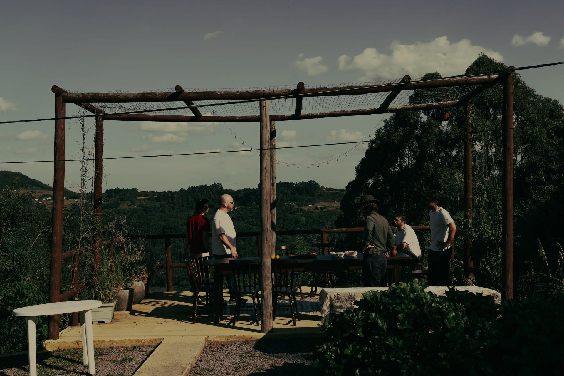 Os homens de pé no deck de madeira, com a mesa posta e a serra ao fundo.
