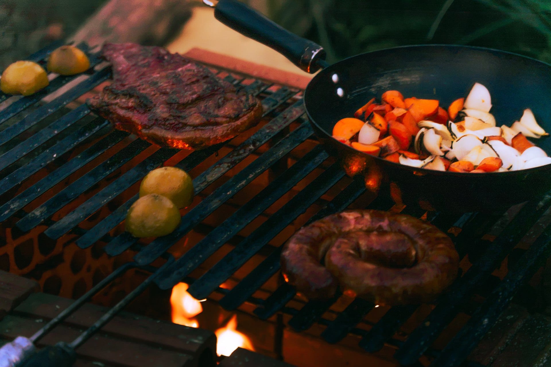 A grelha sobre a brasa, com a carne, a linguiça e a panela de legumes no fogo.