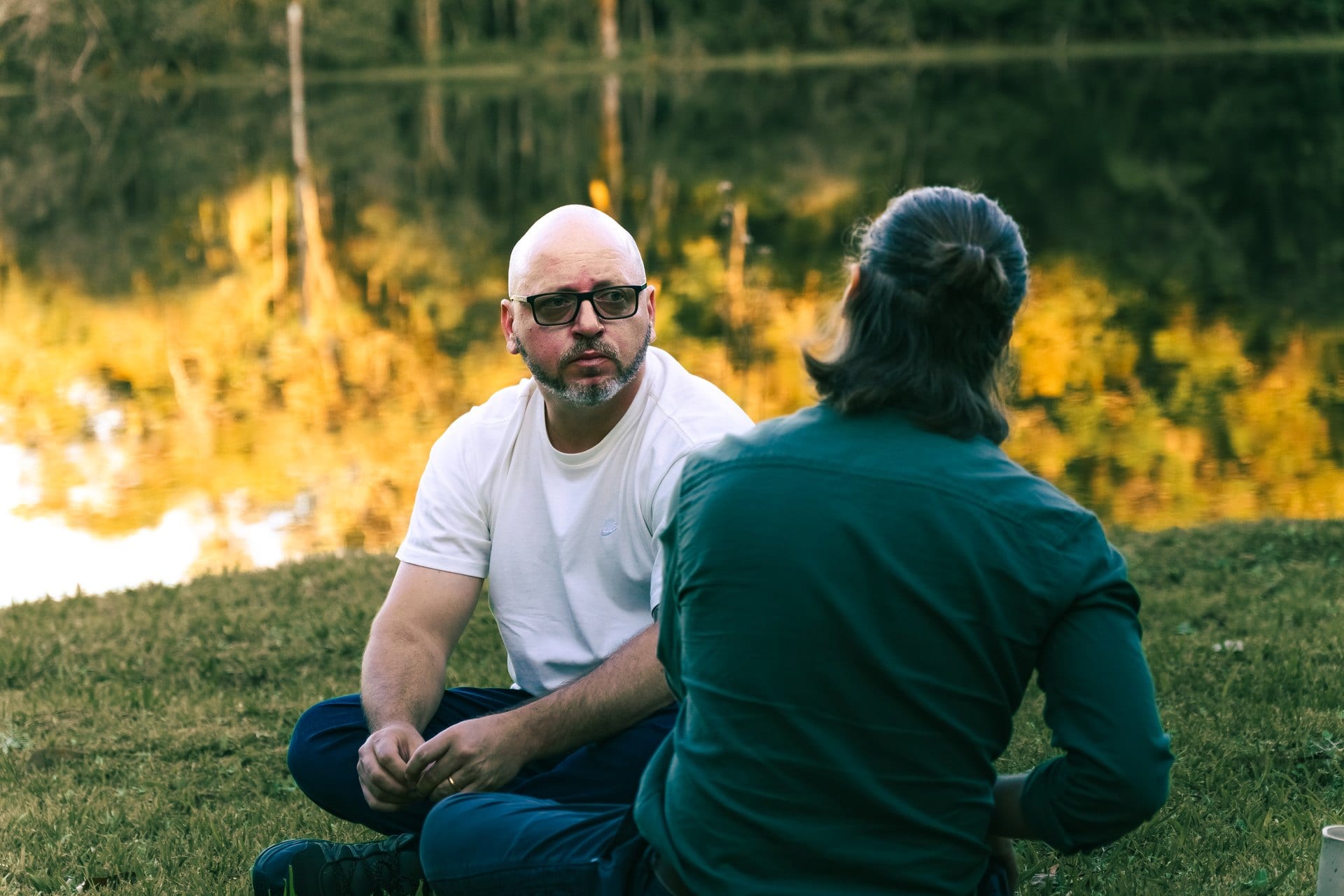 O Aurélio, de óculos e camiseta branca, conversando com o Adriano à beira do lago, com o reflexo dourado das árvores na água atrás.