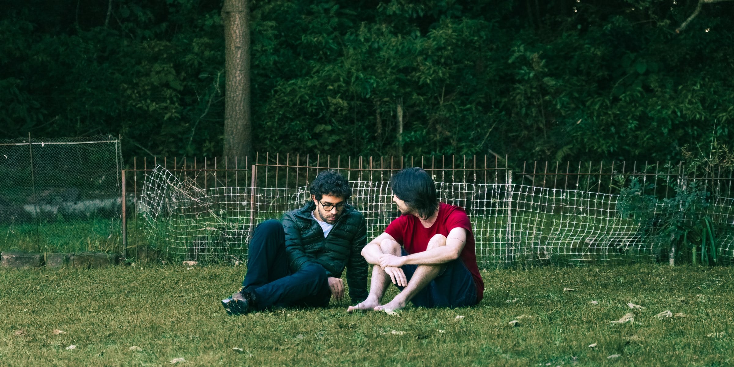 Dois homens da turma de setembro sentados na grama perto da cerca, conversando no fim da tarde.