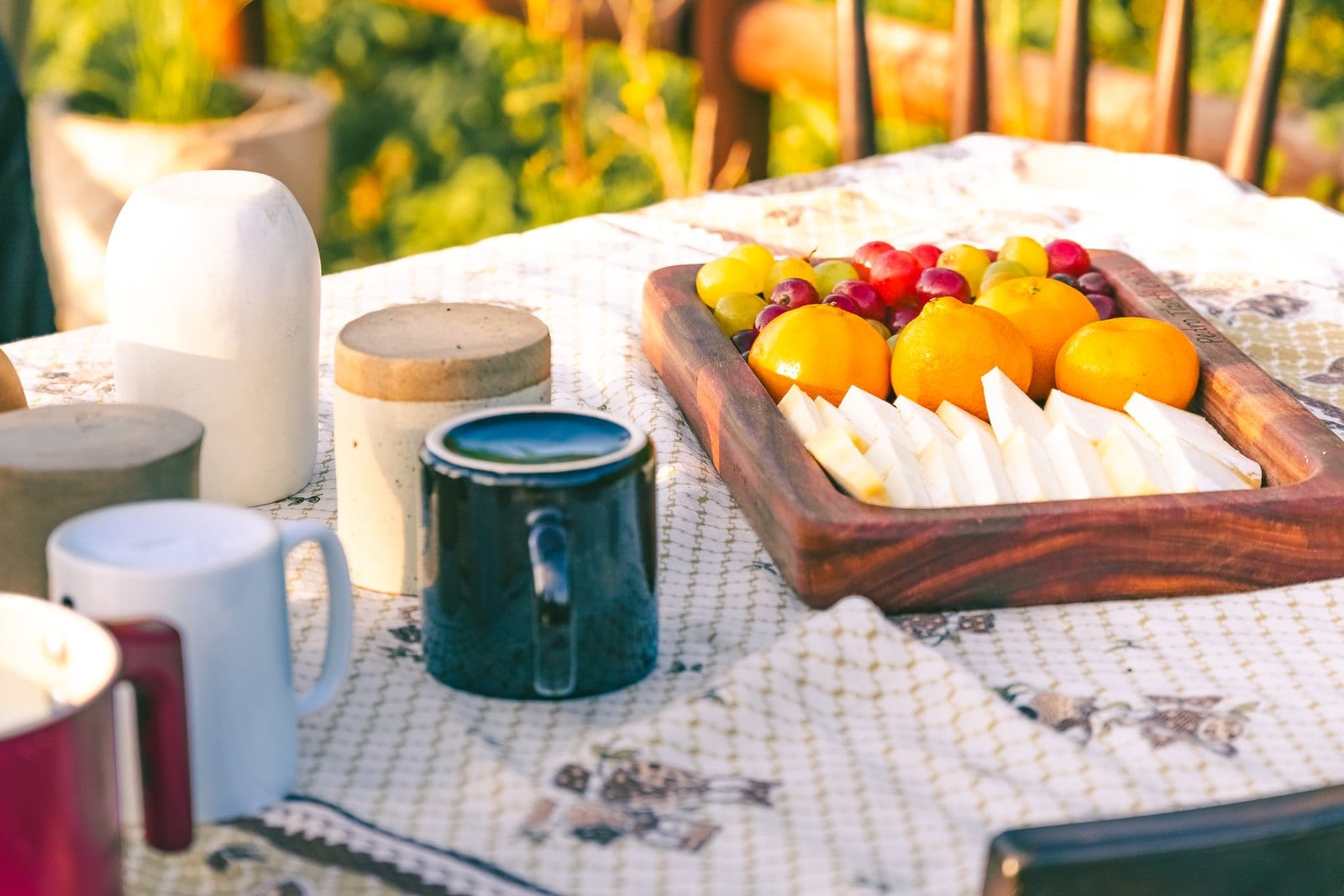 A mesa do café da manhã posta no jardim, com frutas, canecas e a luz da manhã.