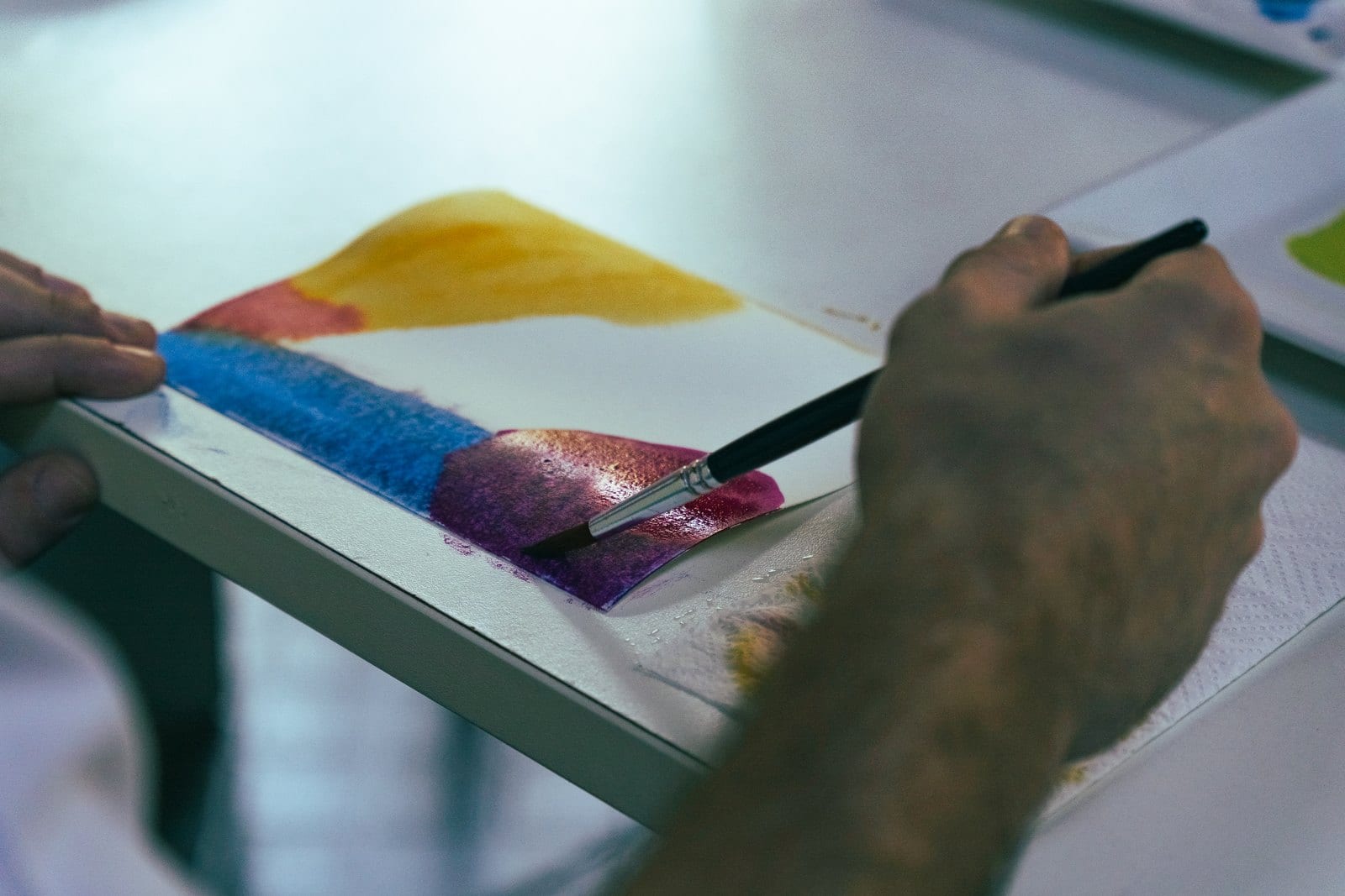 A mão de um homem passando um pincel fino numa aquarela, com faixas de azul, roxo e amarelo sobre o papel branco.