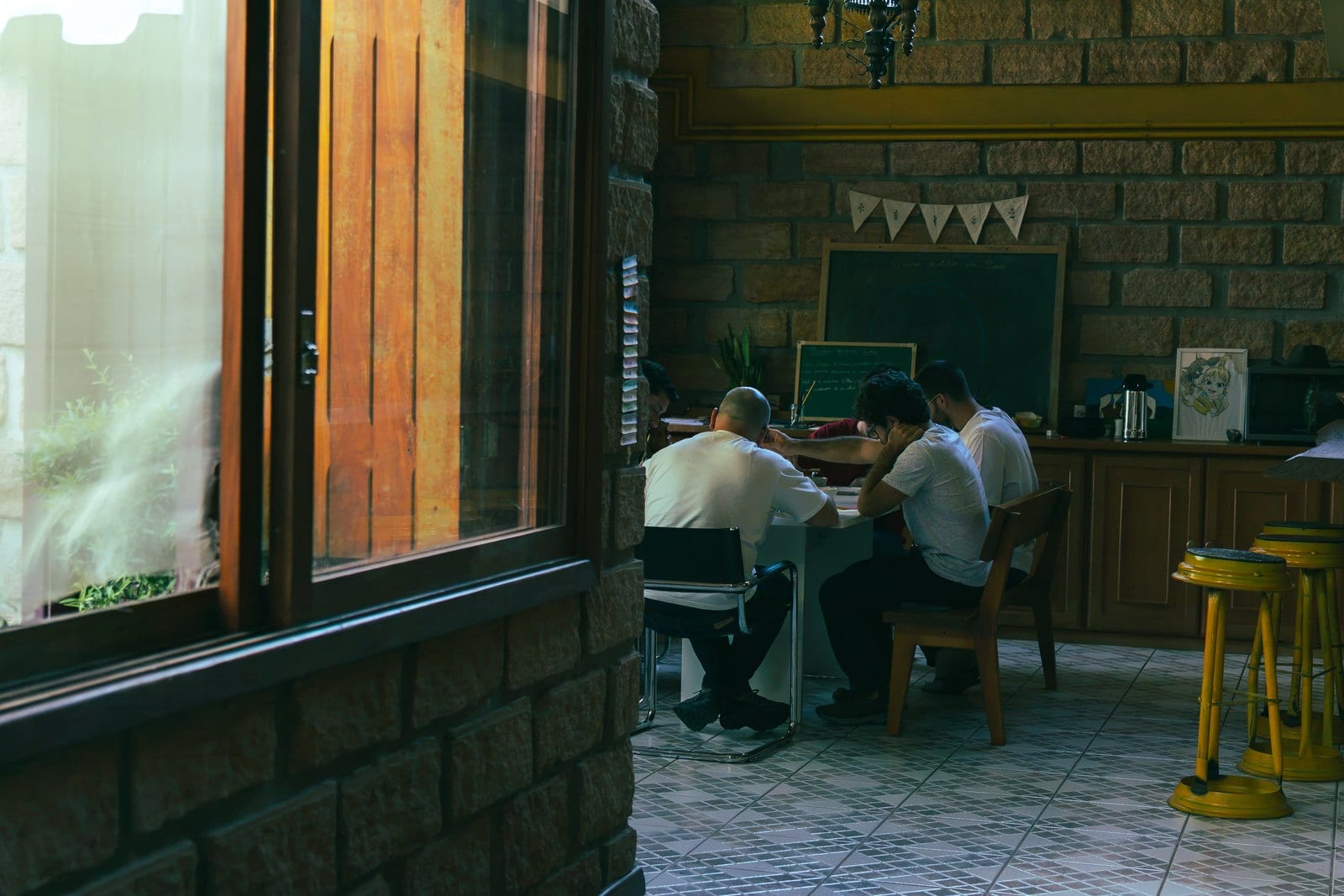 Os homens trabalhando em silêncio na mesa comprida do salão, perto do quadro-negro.