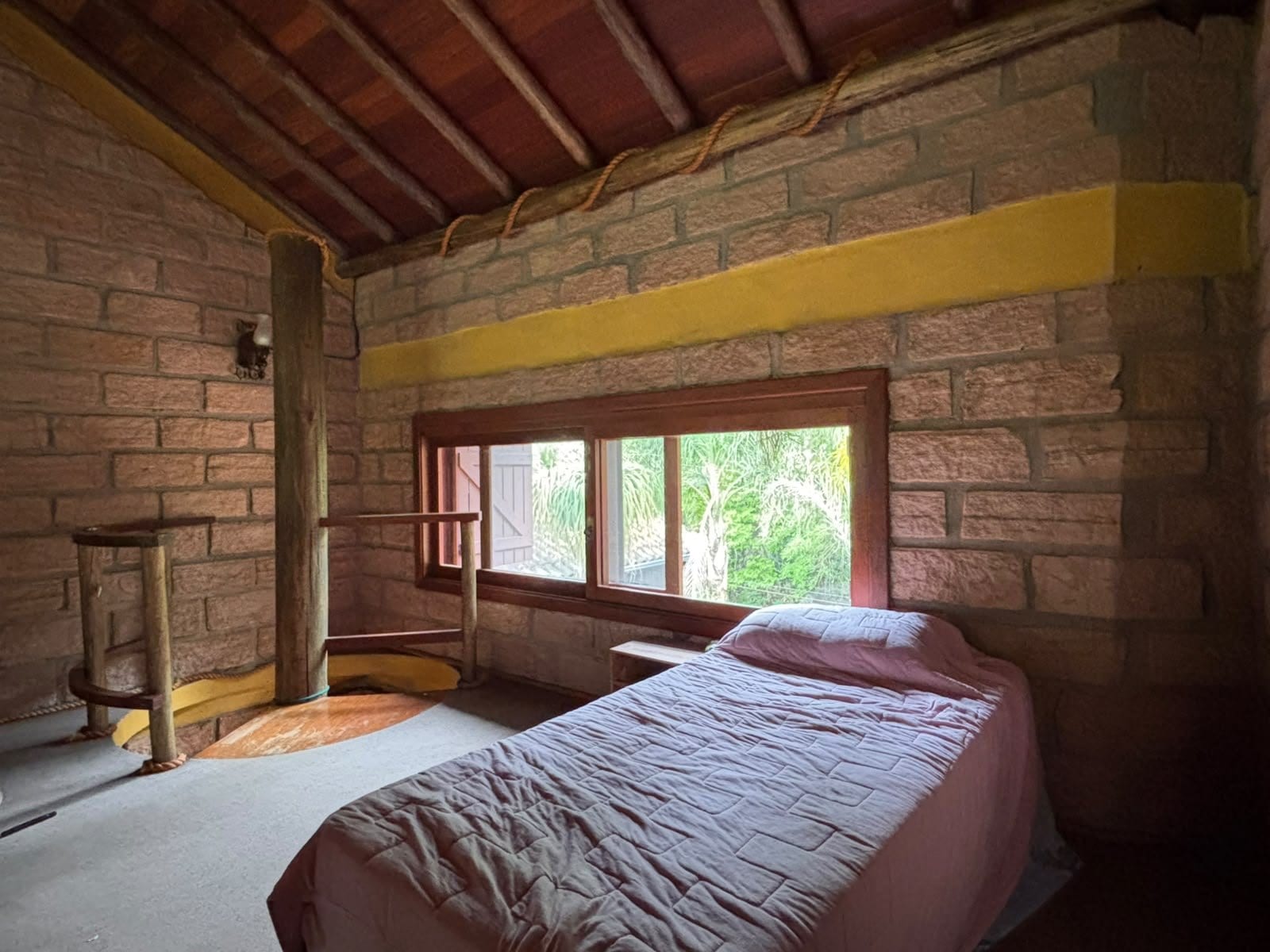 A Estalagem: cama de solteiro sob o telhado de madeira, com a janela comprida aberta pro verde.
