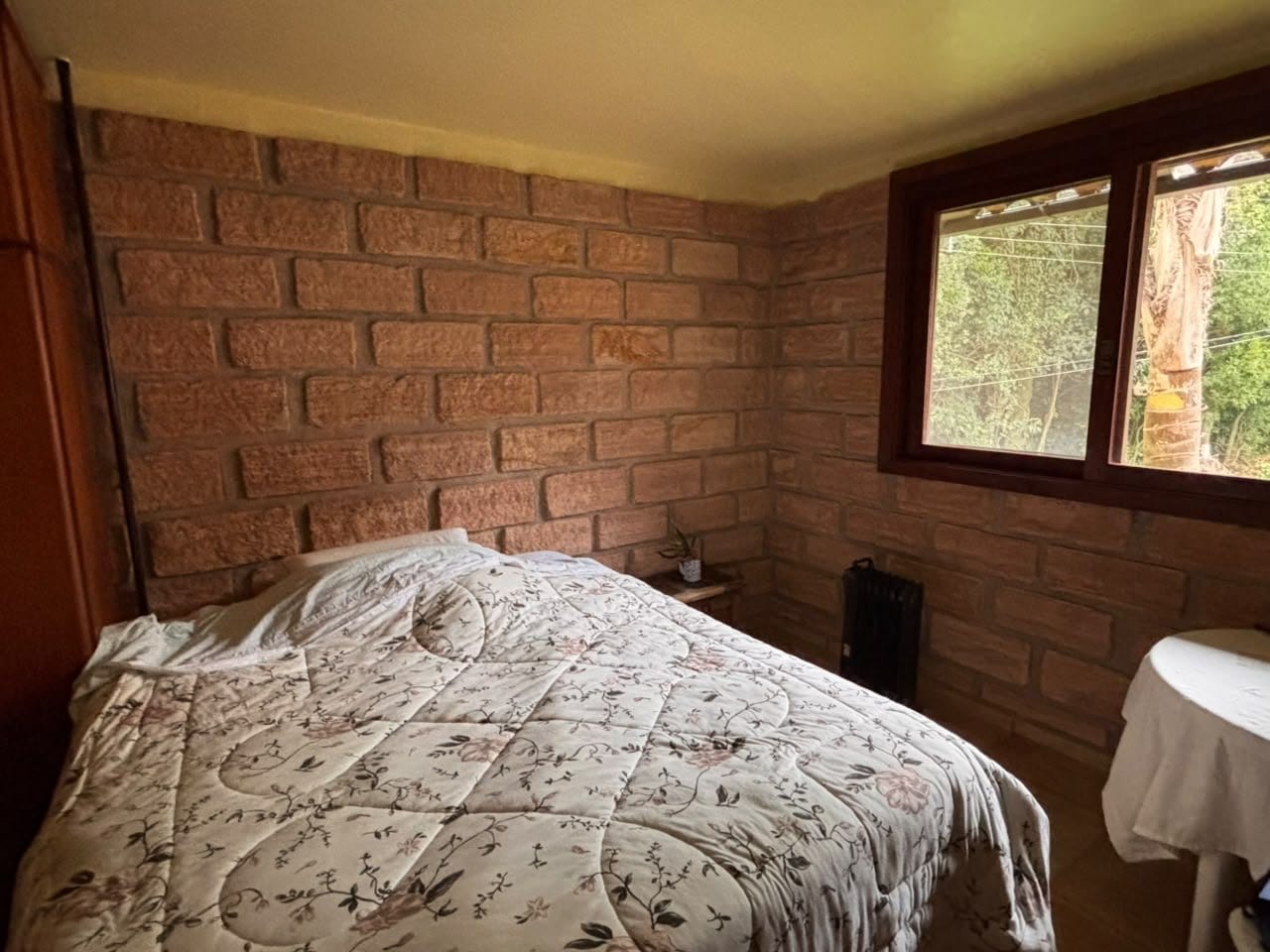 O Quarto da Oliveira: cama de casal queen com colcha florida e parede de tijolo.