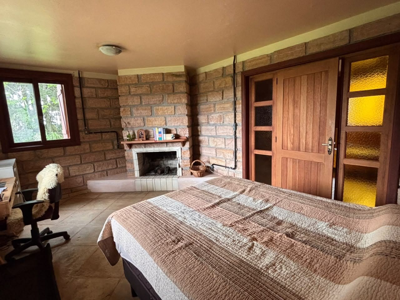 A Torre do Escrivão: cama de casal, a lareira de tijolo no canto e a porta de madeira com vidro que dá pro jardim.