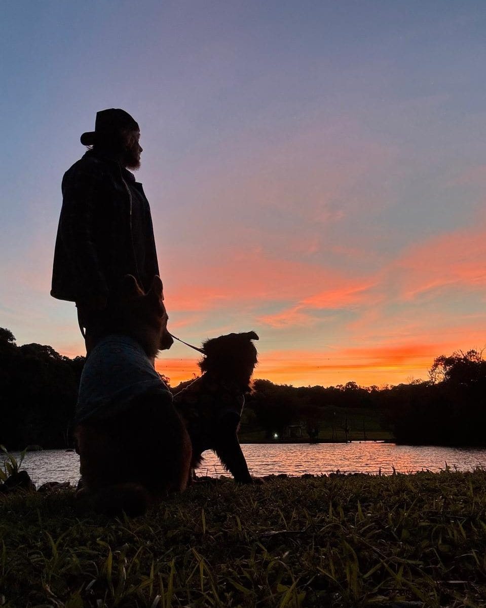 Adriano Rahde de costas com o cachorro, à beira do lago da casa, no fim da tarde.